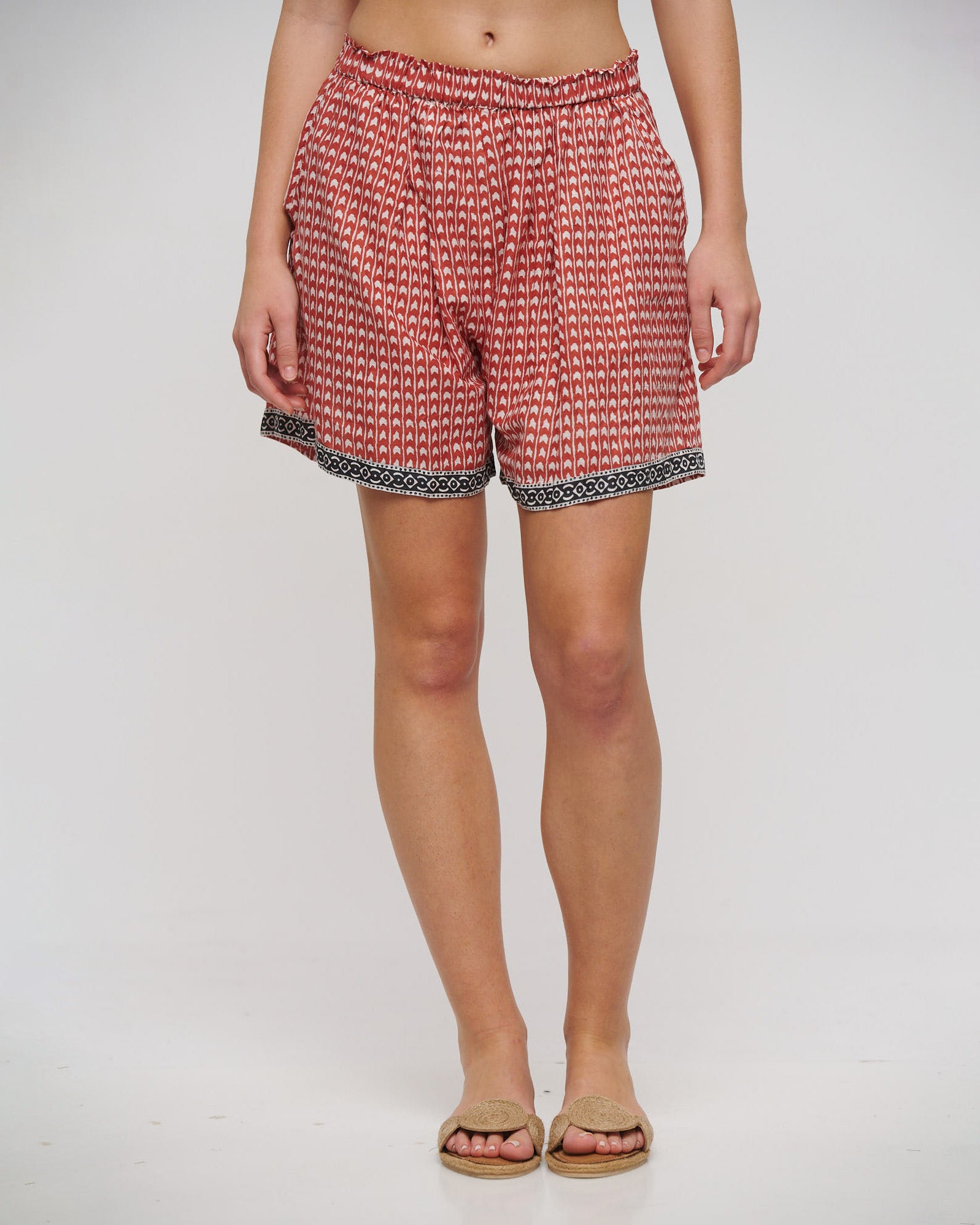 Model trägt locker geschnittene Shorts mit elastischem Bund in Terrakotta und Weiß. Das feine Muster wird durch eine schwarze, ornamentale Bordüre am Saum ergänzt. Kombiniert mit beigen Sandalen.