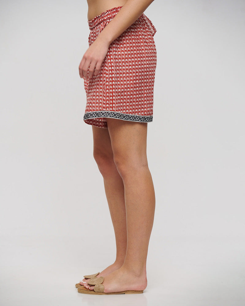 Seitenansicht eines Models in locker fallenden Shorts mit elastischem Bund in Terrakotta und Weiß. Das feine, vertikale Muster wird durch eine schwarze Bordüre mit ornamentalem Design am Saum ergänzt. Kombiniert mit beigen Sandalen.