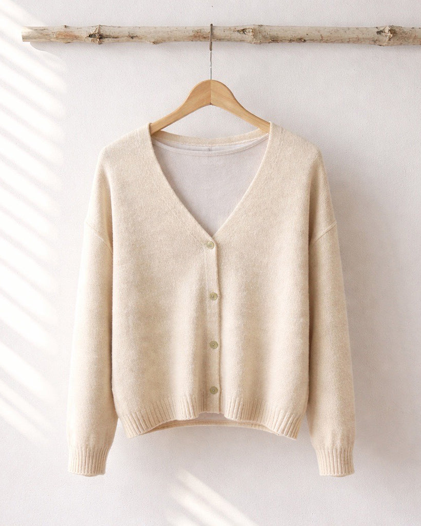 Beiger Cardigan mit V-Ausschnitt und durchgehender Knopfleiste, der an einem Holzkleiderbügel vor einer hellen Wand hängt. Das weiche Strickmaterial und die gerippten Bündchen sind im sanften Tageslicht gut erkennbar.
