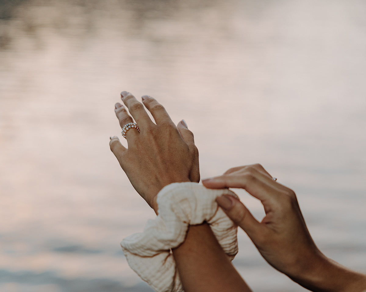 Nahaufnahme zweier Hände vor unscharfem Wasserhintergrund: Eine Hand trägt einen zarten Perlenring, während die andere einen beigen Scrunchie am Handgelenk richtet.