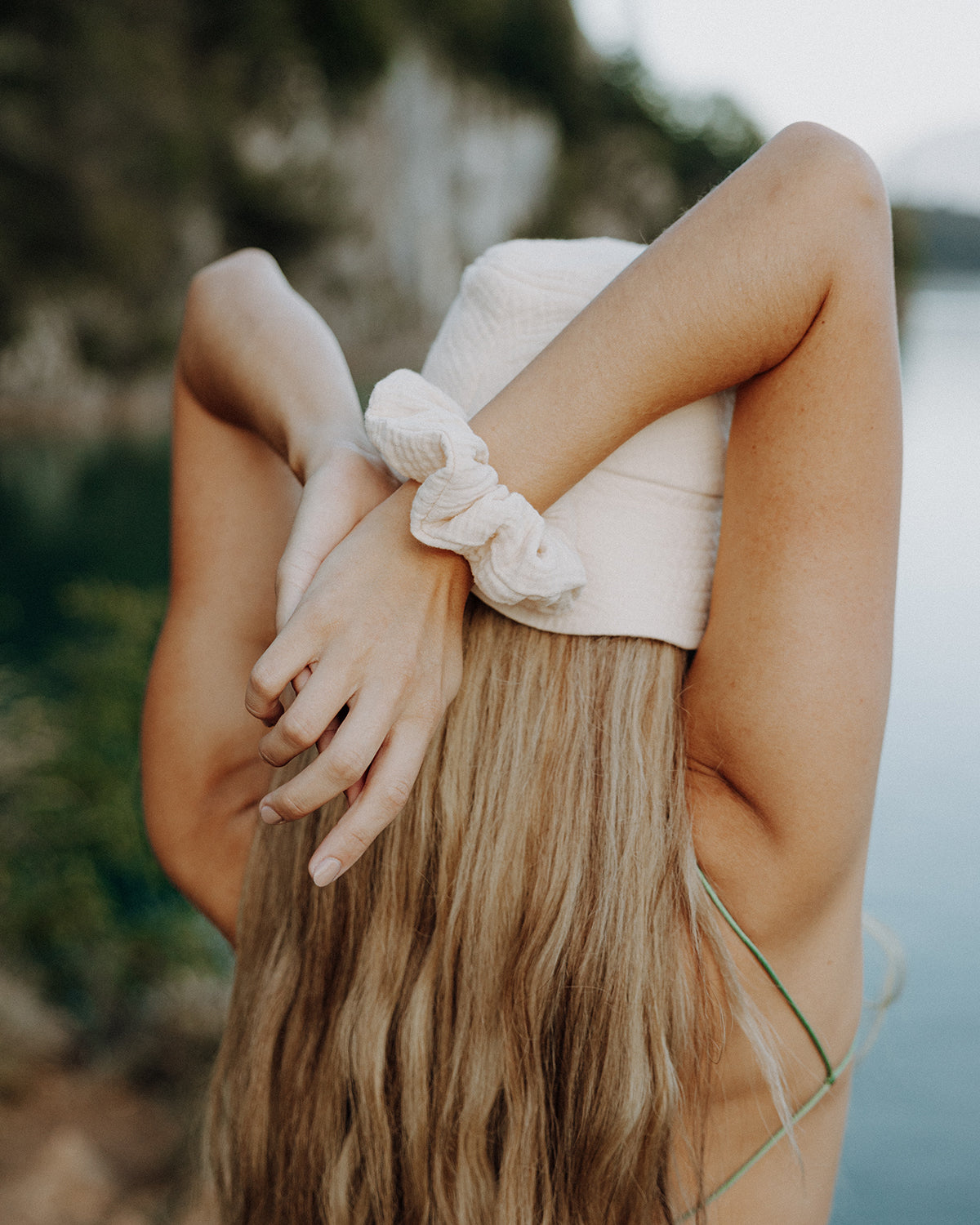 Rückansicht einer Frau mit langen blonden Haaren, die die Arme über dem Kopf verschränkt und einen beigen Scrunchie vor einer natürlichen Fels- und Wasserlandschaft im Hintergrund.
