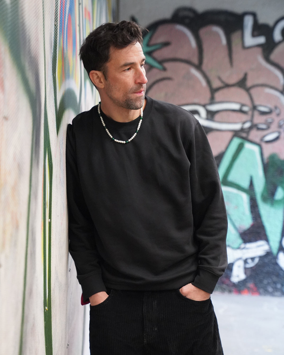 Mann in schwarzem Sweatshirt lehnt an einer Graffiti-Wand und trägt eine Perlenkette für Herren mit weißen Perlen und grünen Akzenten, urbanes Streetstyle-Motiv.