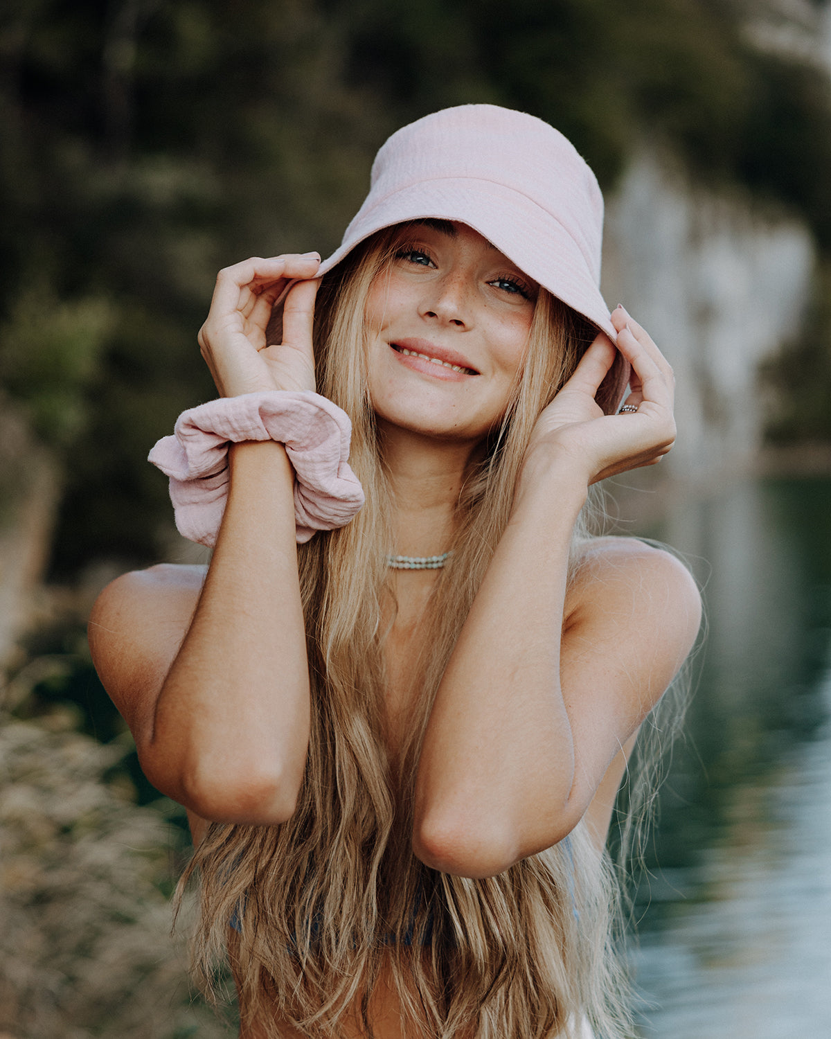 Lächelnde Frau mit langem, blondem Haar trägt einen pastellrosa Bucket Hat und einen pastellrosa Scrunchie am Handgelenk, aufgenommen im Freien vor unscharfem Naturhintergrund am Wa