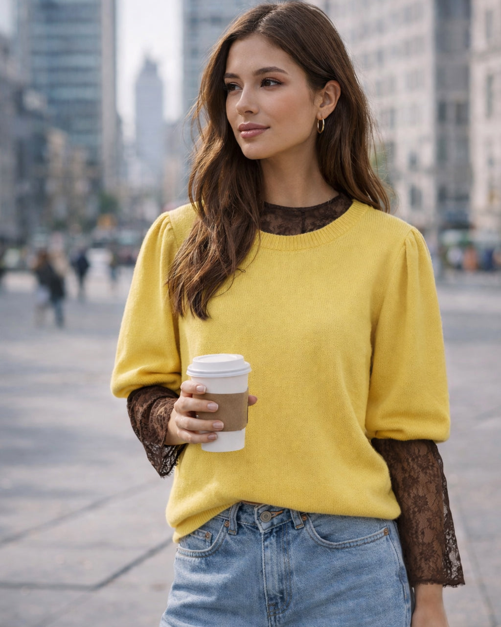 Eine Frau steht in einer Stadtumgebung und hält einen Kaffeebecher in der Hand. Sie trägt einen gelben Pullover mit schwarzen Spitzenärmeln und eine blaue Jeans, im Hintergrund sind unscharfe Hochhäuser zu sehen.