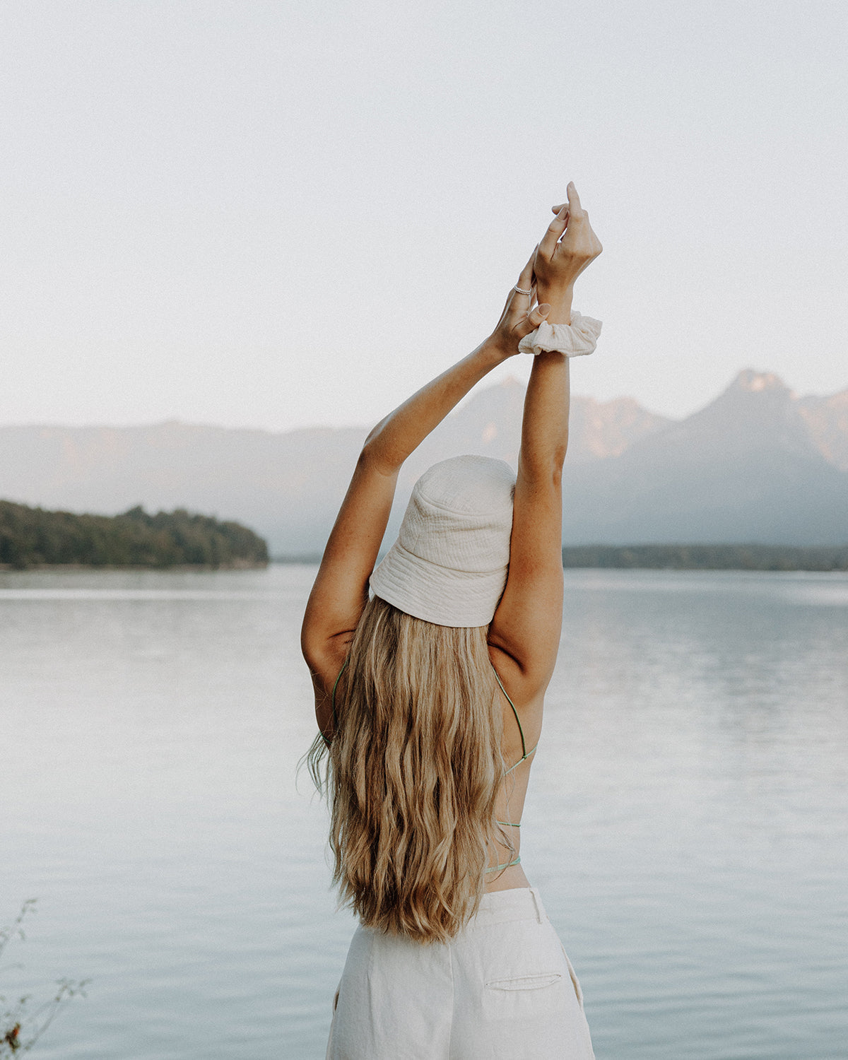 Rückansicht einer Frau mit langen blonden Haaren, die am Seeufer steht und die Arme über dem Kopf streckt. Sie trägt einen beigen Bucket-Hat, einen beigen Scrunchie am Handgelenk, ein grünes Spaghettiträger-Top und eine helle Hose, vor einer ruhigen Berg- und Wasserlandschaft.
