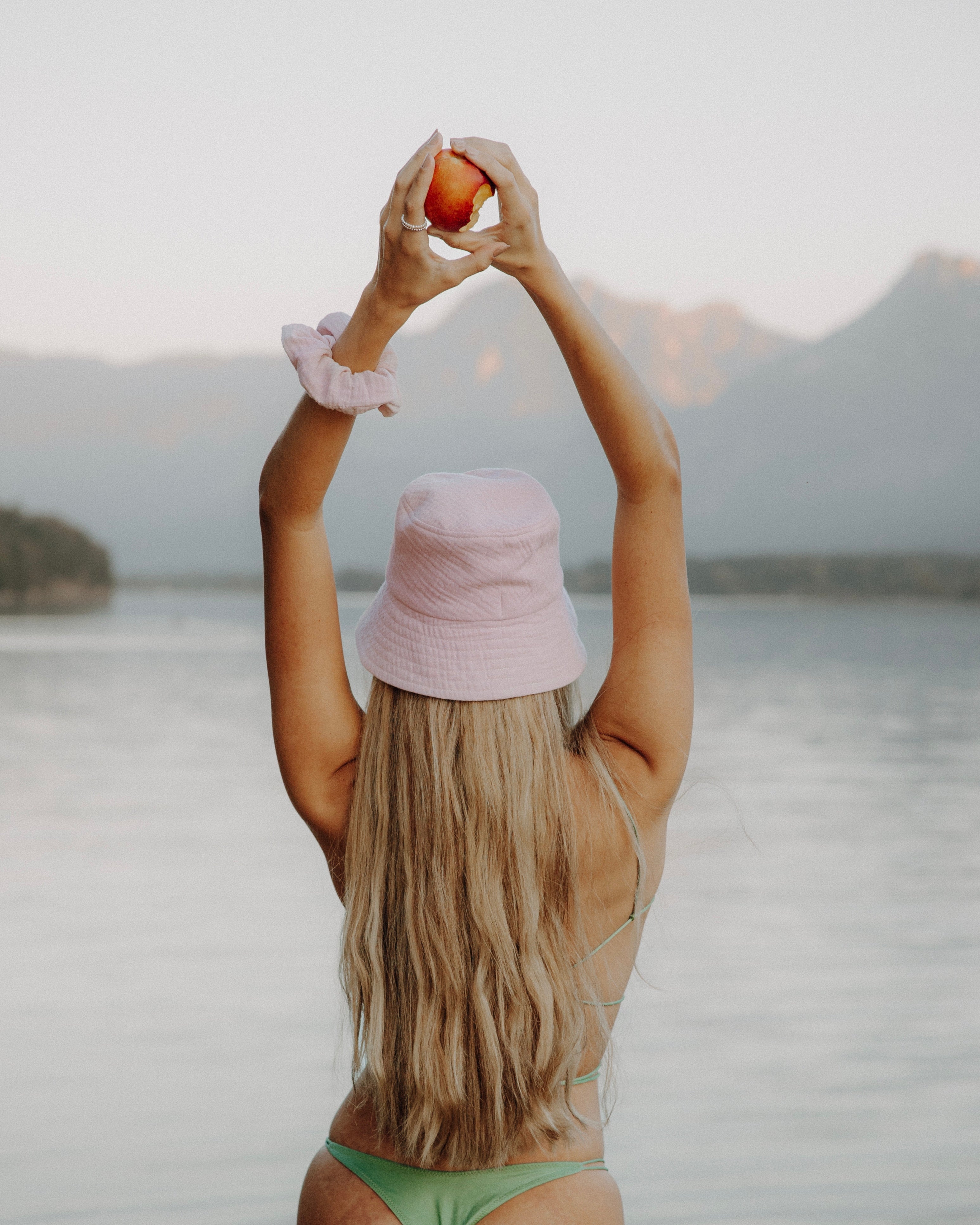 Frau mit langem, blondem Haar trägt einen pastellrosa Bucket Hat und einen pastellrosa Scrunchie am Handgelenk, steht mit erhobenen Armen vor einem türkisfarbenen See und Felslandschaft im Hintergrund.