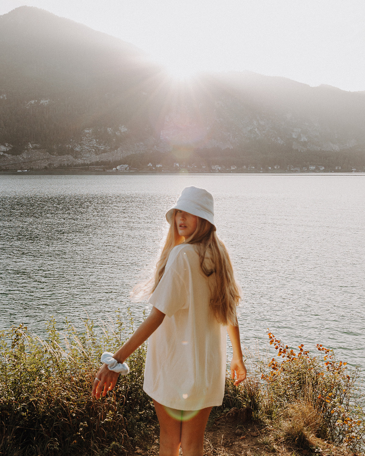 Frau mit langem, blondem Haar trägt einen hellblauen Bucket Hat und einen hellblauen Scrunchie am Handgelenk, dreht sich am Ufer eines Sees vor Bergkulisse im warmen Sonnenlicht um.