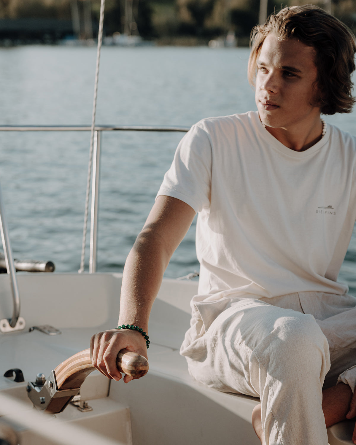 Junger Mann in weißem T-Shirt sitzt auf einem Segelboot, hält das Holzruder und trägt ein grünes Perlenarmband am Handgelenk, im Hintergrund glitzernder See.