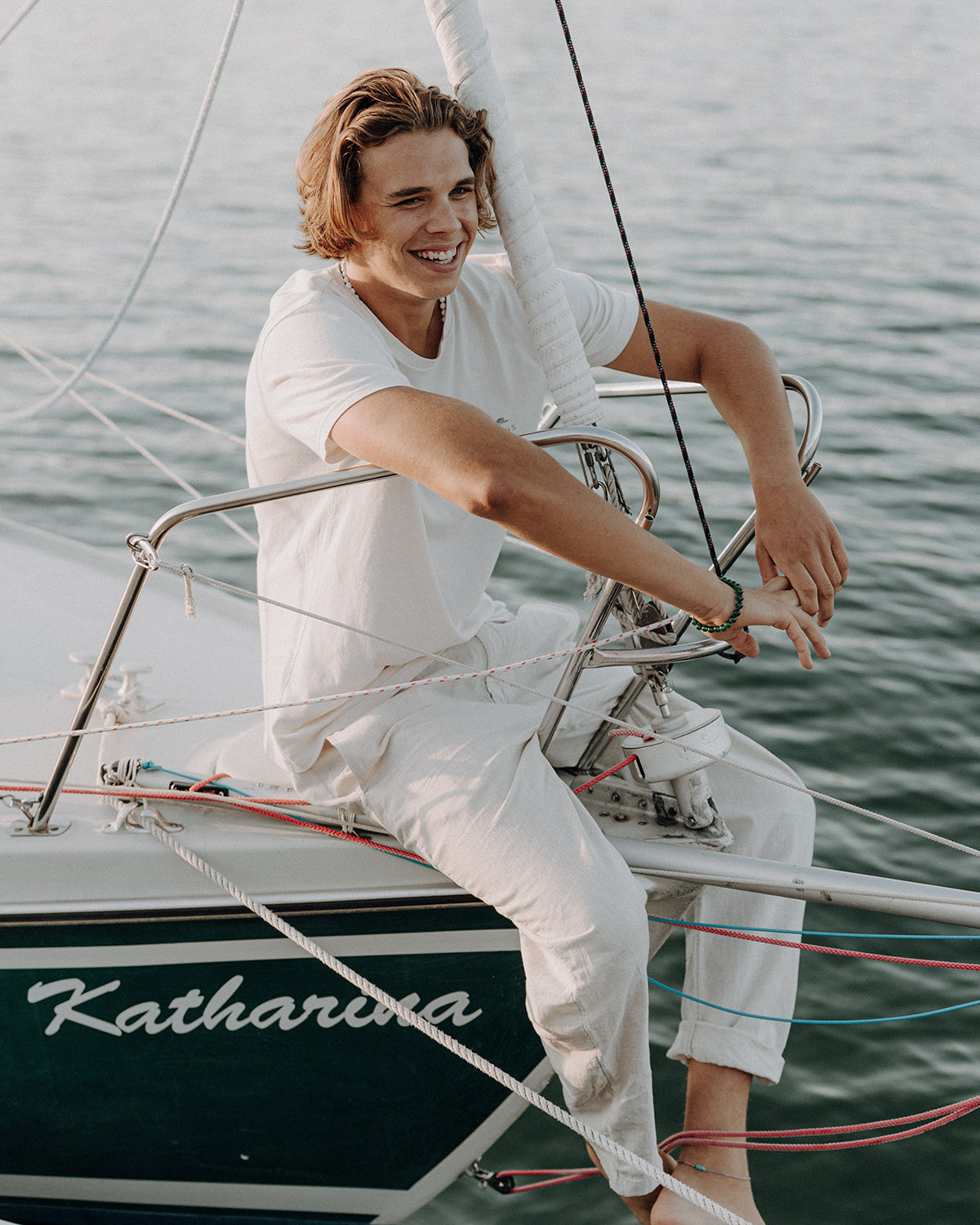 Lächelnder junger Mann in weißer Kleidung sitzt auf einem Segelboot am See und trägt ein grünes Perlenarmband am Handgelenk.