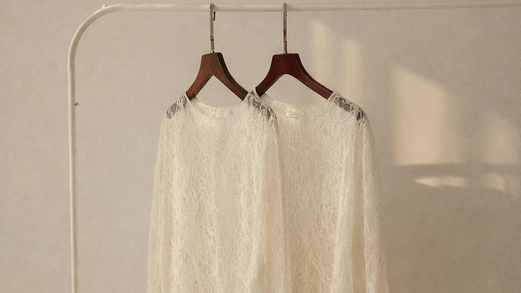 Zwei cremefarbene Spitzenblusen hängen auf Holzkleiderbügeln an einer weißen Kleiderstange. Warmes Sonnenlicht fällt von der Seite auf die Stoffe und wirft weiche Schatten an die Wand im Hintergrund.