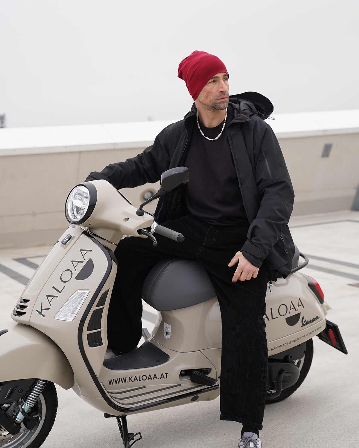 Stilvoller Mann mit roter Mütze sitzt auf einem beigen Roller und trägt eine Perlenkette für Herren mit weißen Perlen, moderner urbaner Look.