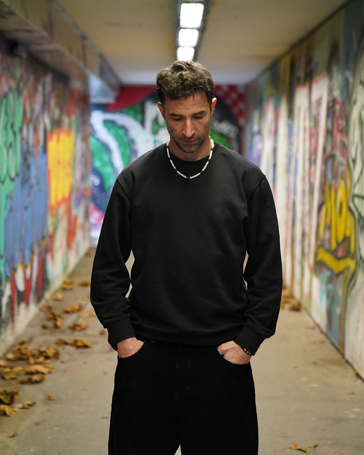 Mann in schwarzem Sweatshirt steht in einem Graffiti-Tunnel und trägt eine Perlenkette für Herren mit weißen Perlen und grünen Akzenten, urbaner und moderner Streetstyle.