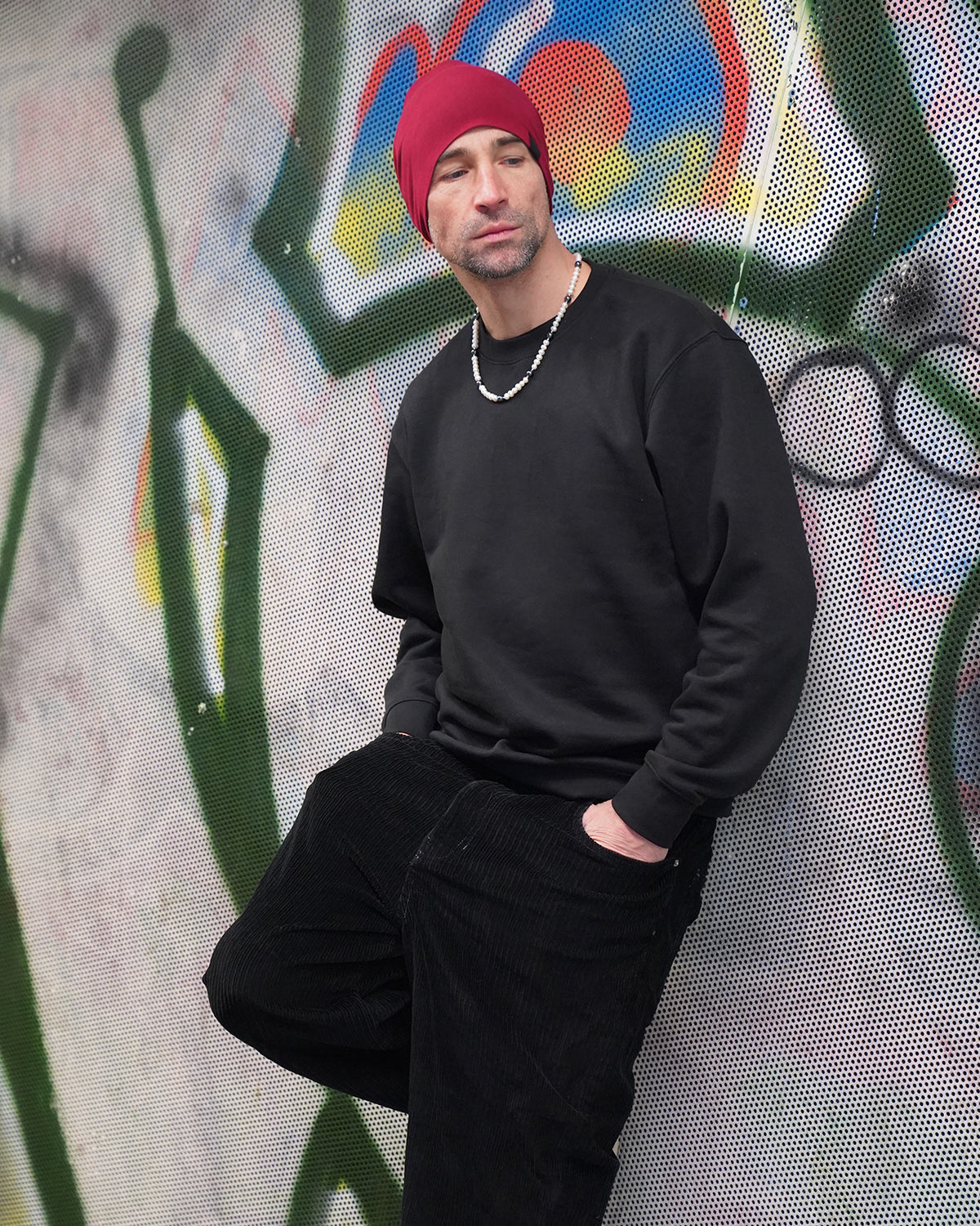 Mann mit roter Mütze lehnt an einer Graffiti-Wand und trägt eine moderne Perlenkette für Herren mit weißen Perlen, urbaner Streetstyle in Schwarz