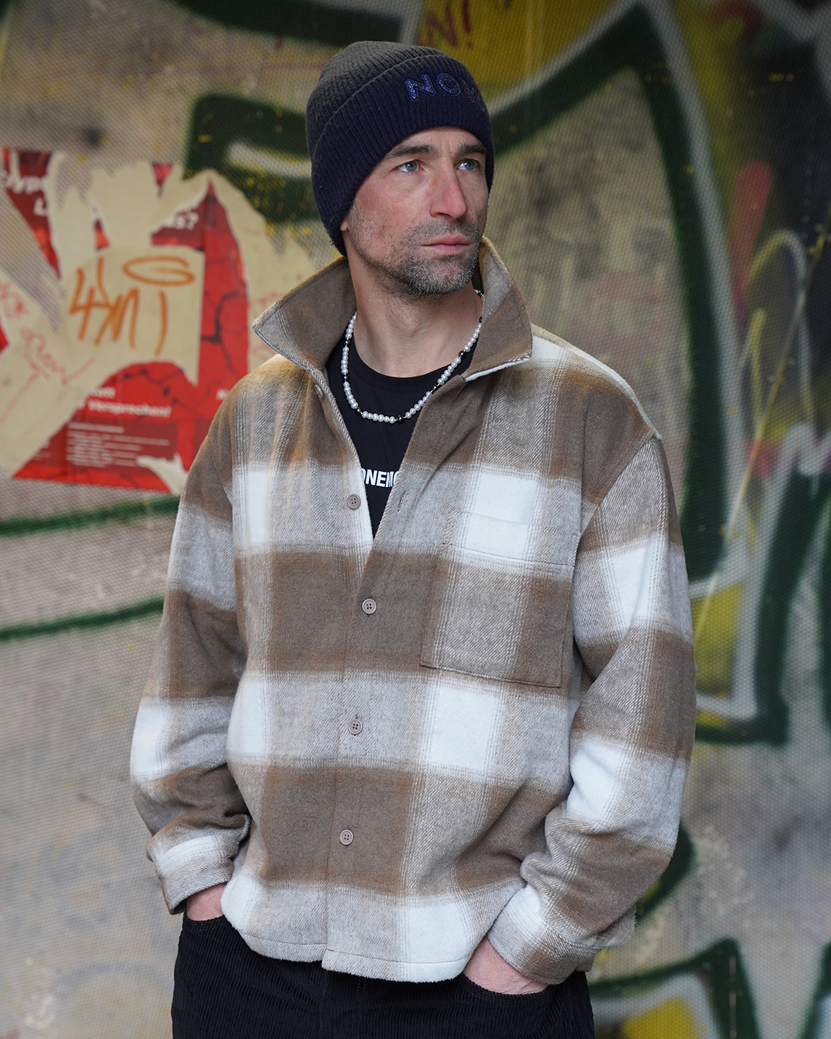 Mann mit dunkler Mütze und karierter Jacke steht vor einer Graffiti-Wand und trägt eine Perlenkette für Herren aus weißen Perlen, urbaner Streetstyle-Look.