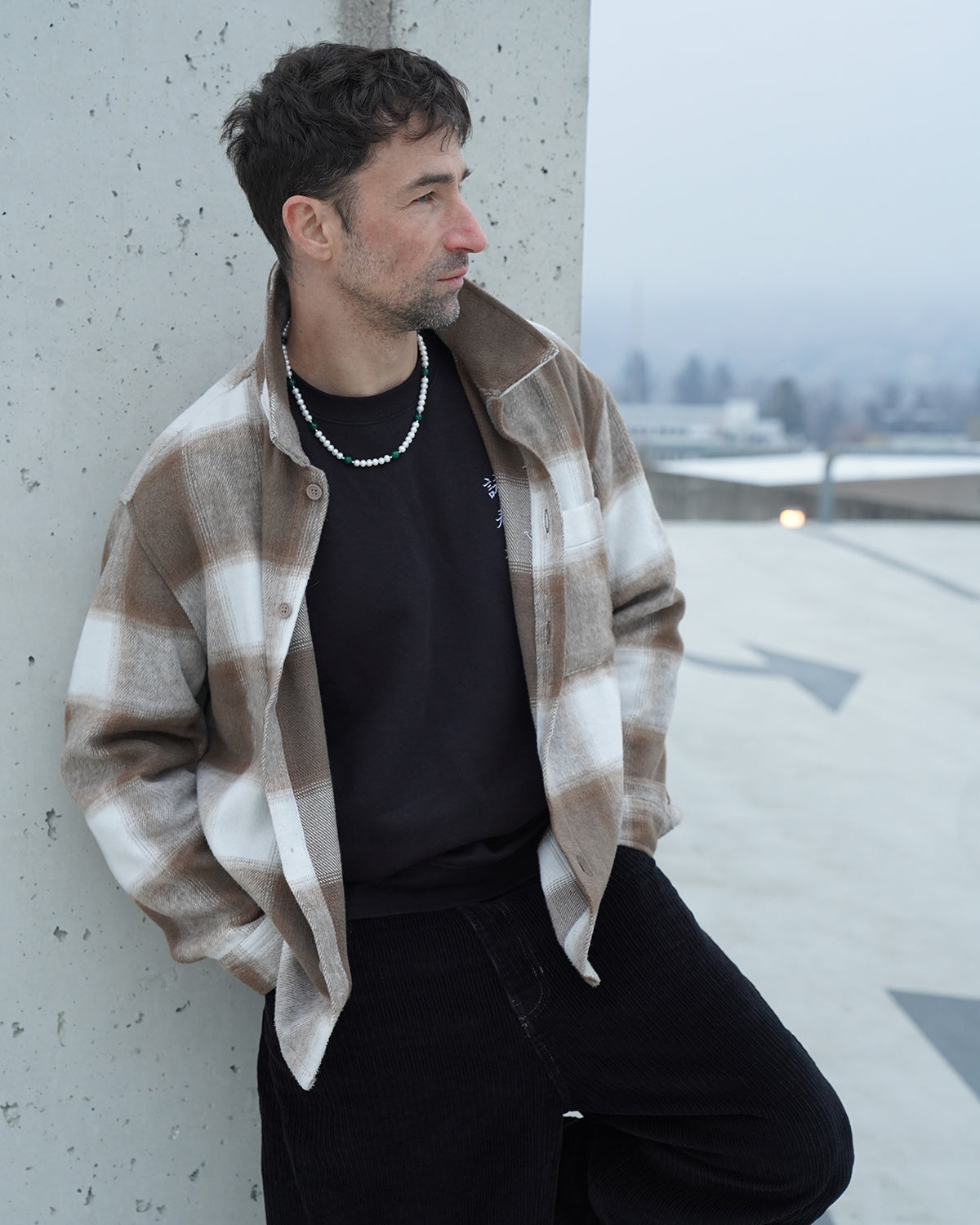 Mann mit karierter Jacke lehnt an einer Betonwand im urbanen Umfeld und trägt eine Perlenkette für Herren mit weißen Perlen und grünen Akzenten, moderner Streetstyle-Look.