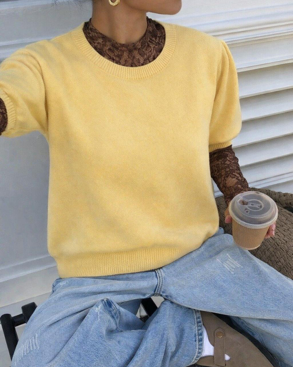 Eine Person sitzt auf einem Stuhl und trägt einen gelben Kurzarm-Strickpullover über einem braunen Spitzentop mit langen Ärmeln. Dazu kombiniert sie eine hellblaue Jeans und goldene Ohrringe; in der Hand hält sie einen Kaffeebecher mit Deckel.
