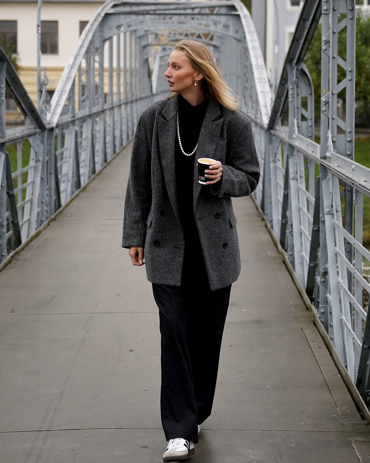 Blonde Frau in schwarzem Outfit mit grauem Mantel und langer Perlenkette geht über eine Metallbrücke; in der Hand hält sie einen Coffee-to-go-Becher und blickt zur Seite.