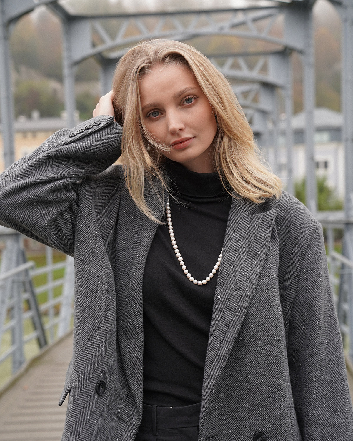 Porträt einer blonden Frau auf einer Metallbrücke, trägt schwarzen Rollkragenpullover und grauen Mantel mit Fischgrätmuster sowie eine lange Perlenkette; sie blickt in die Kamera und fährt sich mit der Hand durch die Haare.