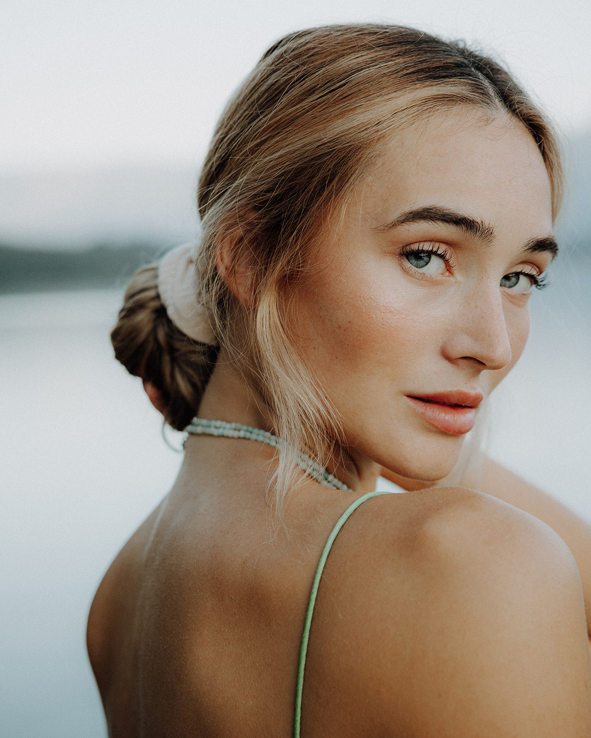 Porträt einer blonden Frau mit lockerem Dutt und beigem Scrunchie, die über die Schulter blickt und eine zarte, mintfarbene Perlenkette zu einem grünen Spaghettiträger-Top trägt, vor ruhiger Wasserlandschaft im Hintergrund.