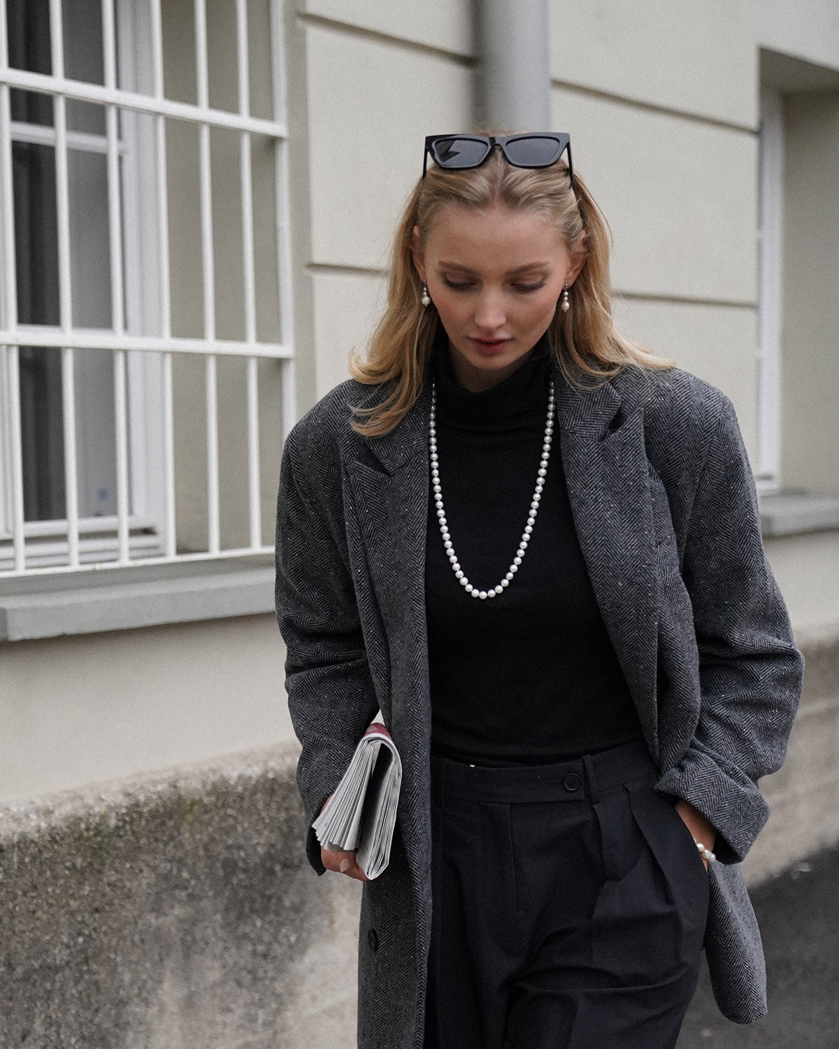 Junge Frau mit blondem Haar vor einem hellen Gebäude, trägt schwarzen Rollkragenpullover, grauen Mantel im Fischgrätmuster und eine lange Perlenkette; eine Sonnenbrille sitzt auf ihrem Kopf, in der Hand hält sie eine gefaltete Zeitung.