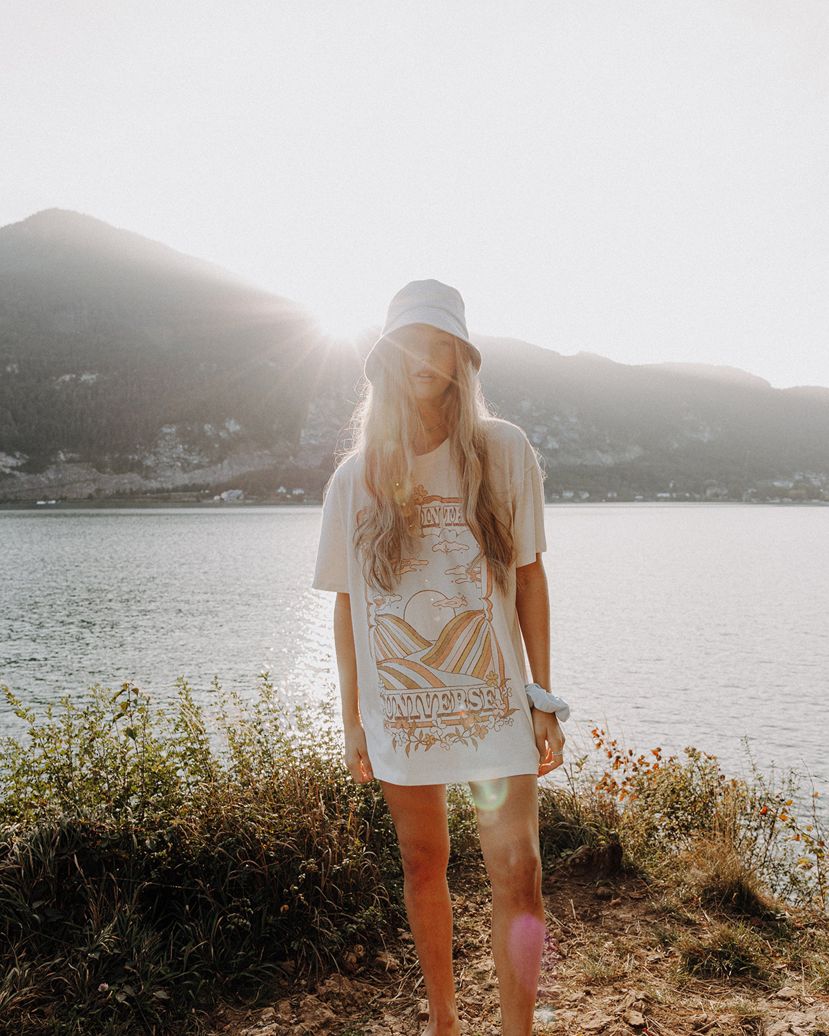 Frau mit langem, blondem Haar trägt einen hellblauen Bucket Hat und einen hellblauen Scrunchie am Handgelenk, steht am Seeufer vor Berglandschaft im warmen Sonnenuntergang.