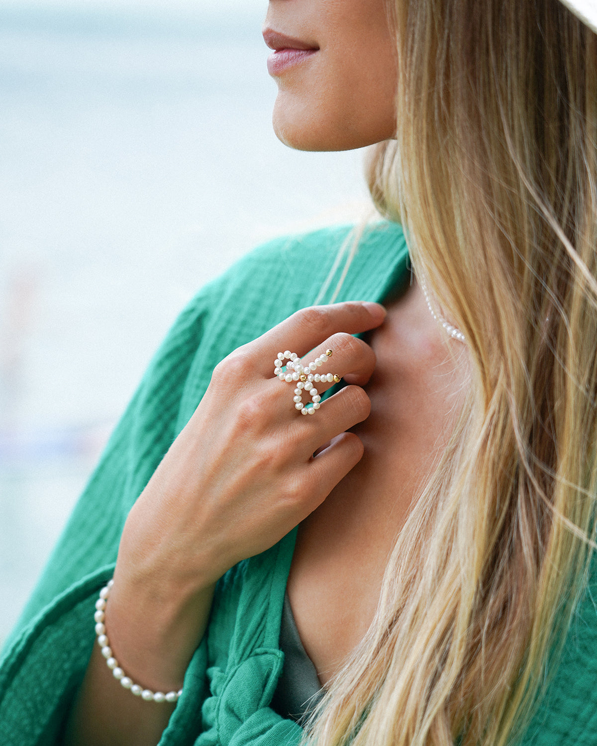 Nahaufnahme einer Frau mit grünem Oberteil, die einen auffälligen Perlenring in Schleifenform sowie ein zartes Perlenarmband trägt und mit der Hand ihr Haar berührt