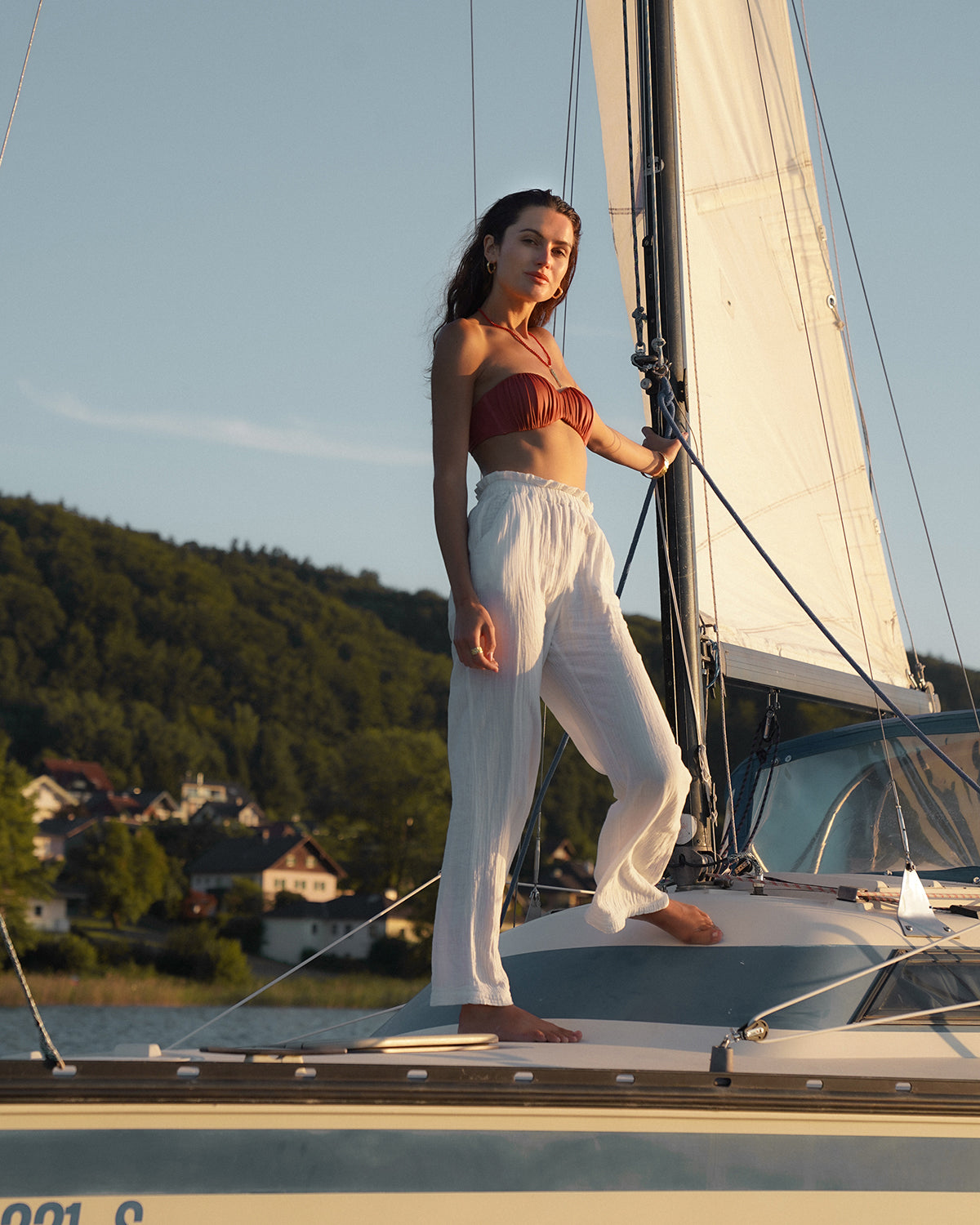 eine Frau mit dunklen Haaren steht auf einem Segelboot und haltet sich fest. Sie trägt einen auffällige Rote Perlenkette mit goldfarbenem Fisch-Anhänger und einen Goldarmreif. Im Hintergrund sieht man etwas vom Meer und eine schöne Landschaft.