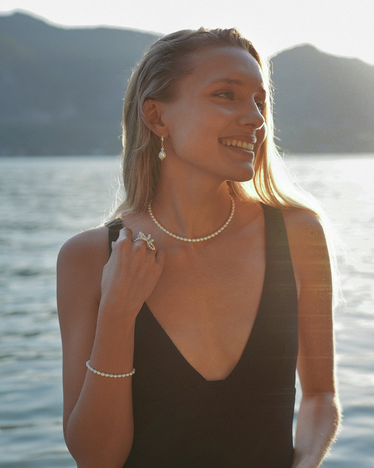 Lächelnde Frau im warmen Abendlicht am Meer, trägt ein schwarzes Kleid sowie eine feine Perlenkette, passende Perlenohrringe und ein Perlenarmband; im Hintergrund sind Wasser und Berge zu sehen.