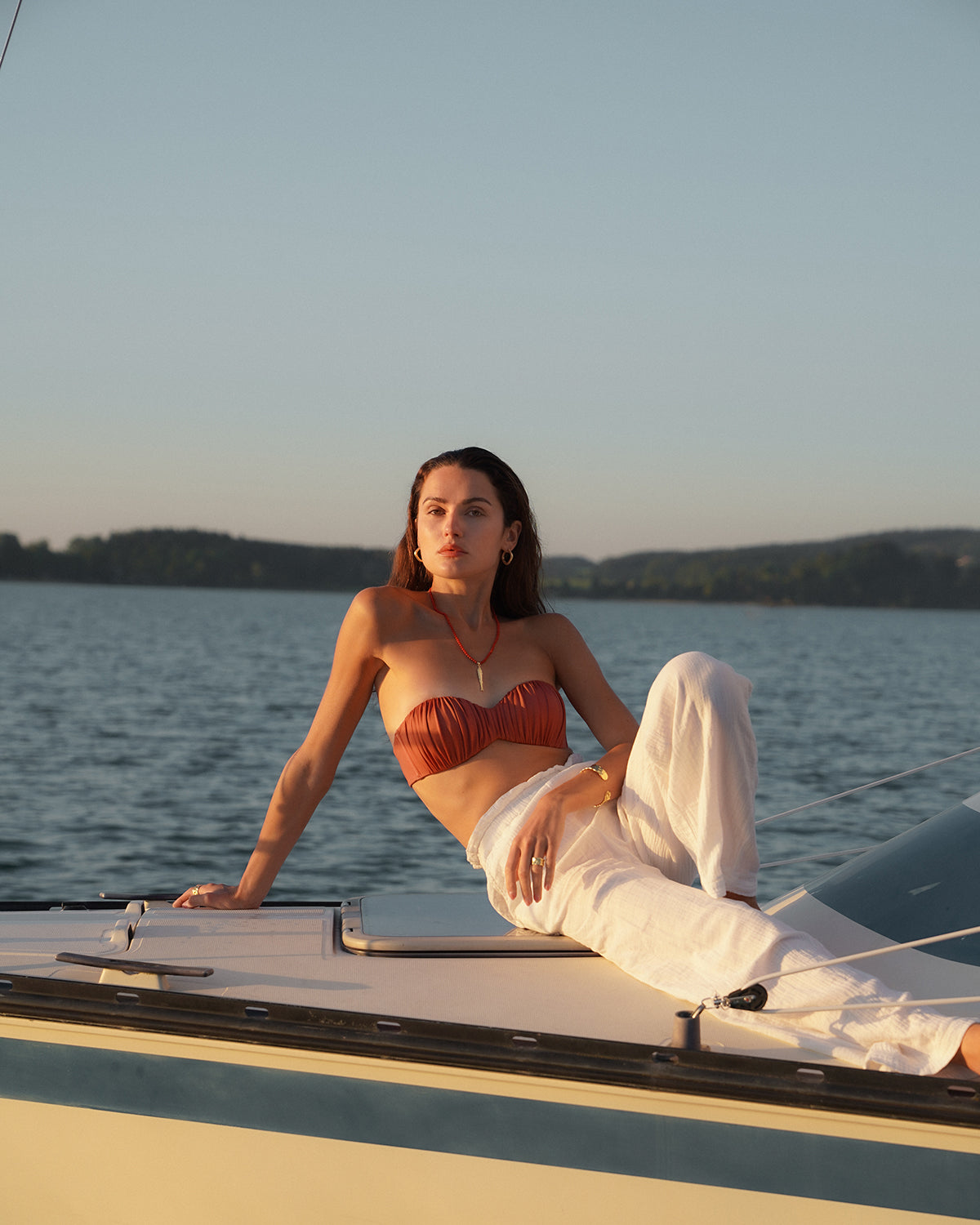 Eine Frau liegt entspannt auf dem Deck eines Segelboots vor einer ruhigen Wasserlandschaft bei Sonnenuntergang. Sie trägt ein rostfarbenes Bandeau-Top und eine weite weiße Hose, dazu eine rote lange Perlenhalskette mit Gold Anhänger und goldenen Schmuck. Im Hintergrund sind ein See und bewaldete Hügel zu sehen, während warmes Abendlicht die Szene beleuchtet.