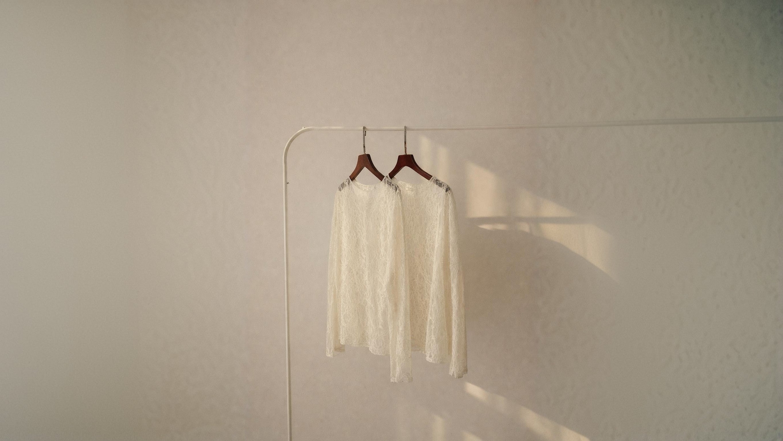 Zwei cremefarbene, langärmelige Spitzenblusen hängen auf dunklen Holzkleiderbügeln an einer schlichten weißen Kleiderstange vor einer hellen Wand. Warmes Sonnenlicht fällt von der Seite ein und wirft weiche Schatten auf die Wand.