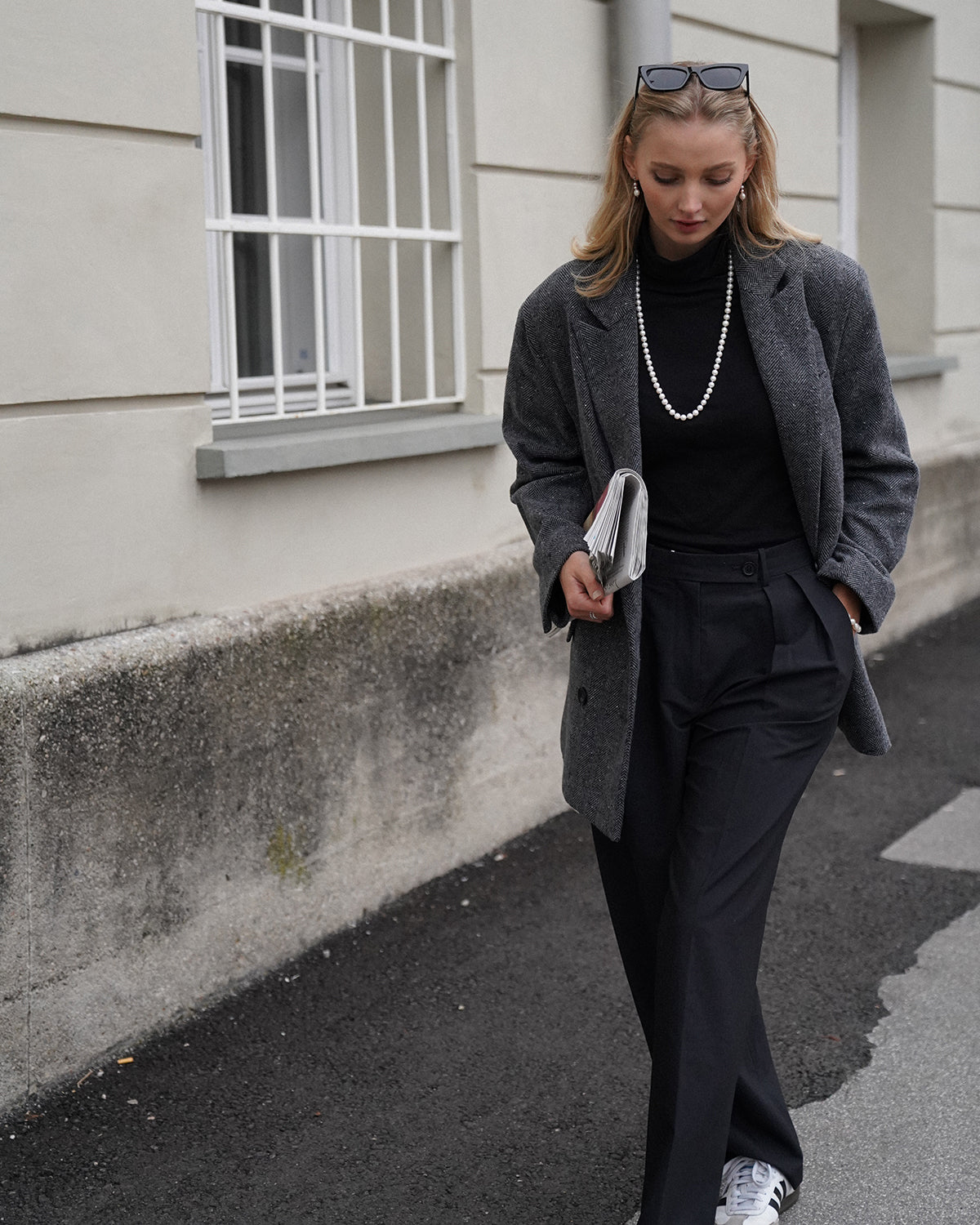 Junge Frau mit blondem Haar vor einem hellen Gebäude, trägt schwarzen Rollkragenpullover, grauen Mantel im Fischgrätmuster und eine lange Perlenkette; eine Sonnenbrille sitzt auf ihrem Kopf, in der Hand hält sie eine gefaltete Zeitung.