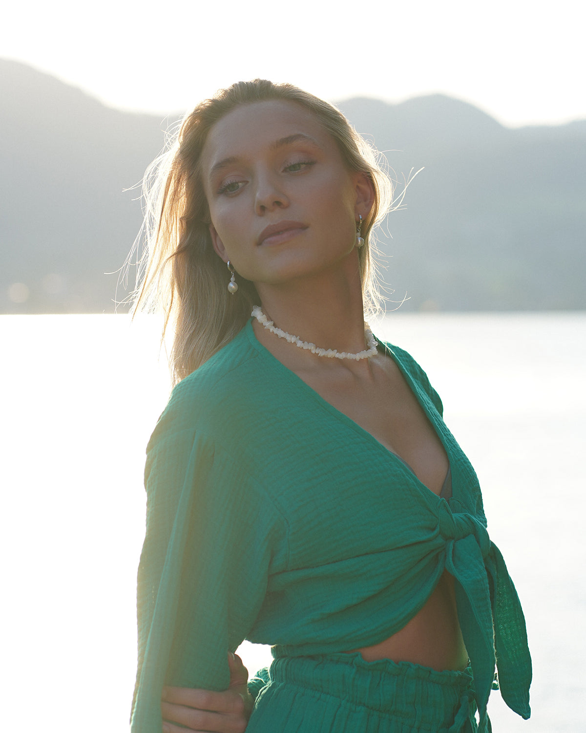 Blonde Frau im grünen Zweiteiler trägt eine weiße Mondstein-Halskette und passende Ohrringe, fotografiert im warmen Abendlicht vor Berg- und Seelandschaft im Slow-Living-Stil.