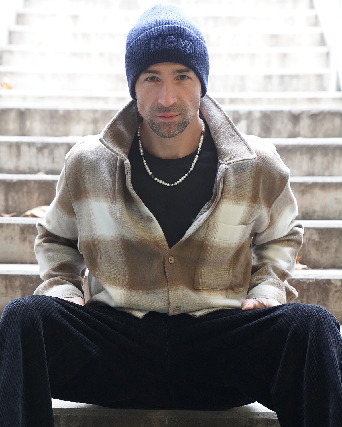Mann mit blauer Mütze sitzt auf einer Treppe und trägt eine stilvolle Perlenkette für Herren mit weißen Perlen, lässiger und moderner Herbstlook.