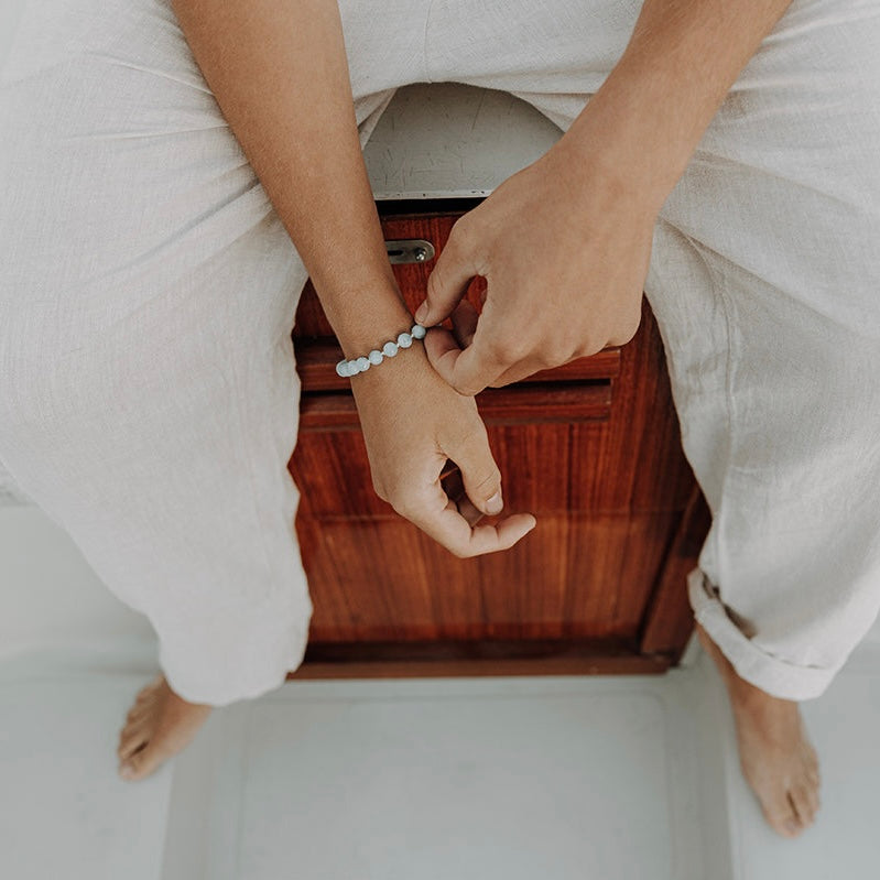 Draufsicht auf eine sitzende Person in weißer Kleidung, die barfuß auf einem Holzkasten sitzt und ein hellblaues Perlenarmband für Herren am Handgelenk zurechtrückt.