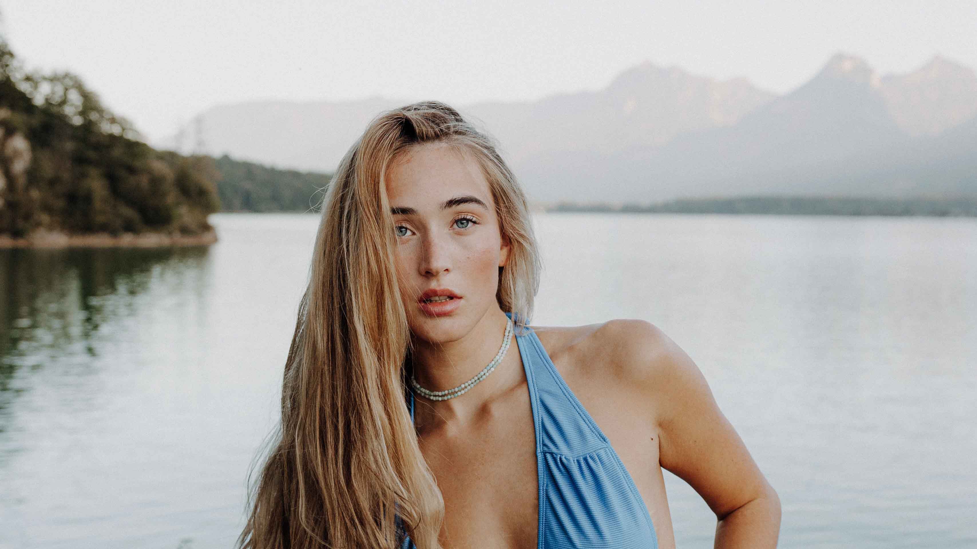 blonde person mit langen Haaren und blauen Top und 2 blaue Halsketten um den Hals. Im Hintergrund ist ein See.