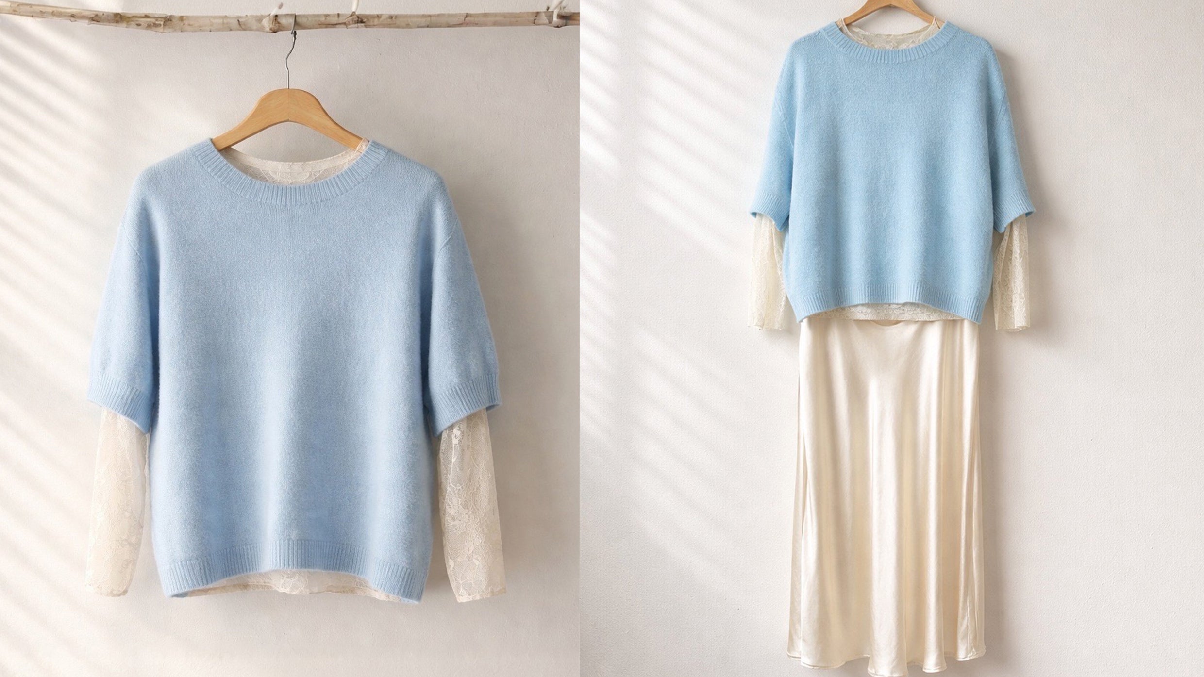 Ein hellblauer, kurzarm Strickpullover hängt auf einem Holzkleiderbügel an einem schlichten Ast vor einer weißen Wand. Darunter ist eine cremefarbene Bluse mit Spitzenärmeln sowie ein langer, fließender Rock in einem sanften Cremeton kombiniert. Weiches Tageslicht fällt von der Seite ein und erzeugt zarte Schatten auf der Wand.