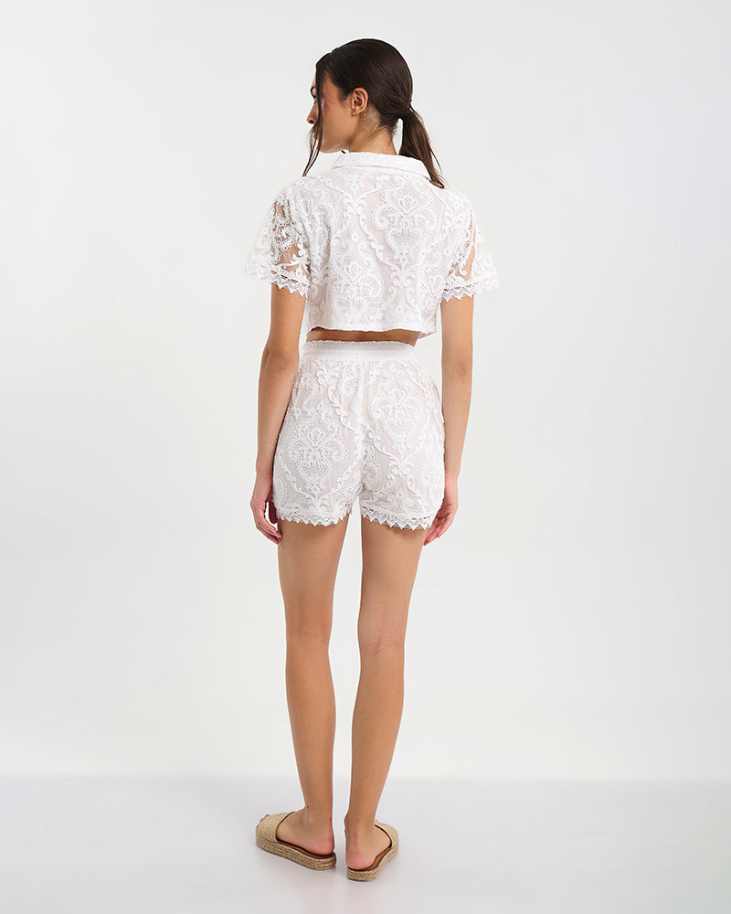 Rückansicht einer Frau in weißem zweiteiligem Spitzen-Set mit kurzärmligem Crop-Top und passender Shorts vor hellem Hintergrund, kombiniert mit flachen Sandalen.