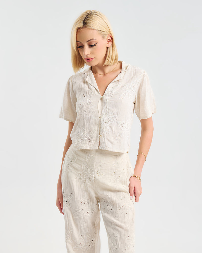 Model trägt ein cremefarbenes zweiteiliges Outfit aus kurzer Bluse mit Knopfleiste und weiter Hose. Beide Teile sind mit floraler Lochstickerei verziert und wirken luftig und leicht. Kombiniert mit dezentem Schmuck.
