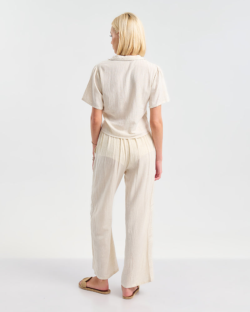 Rückansicht eines Models in einem cremefarbenen zweiteiligen Outfit aus kurzärmeliger Bluse und weiter Hose. Das leichte Material wirkt luftig und wird mit beigen Sandalen kombiniert.