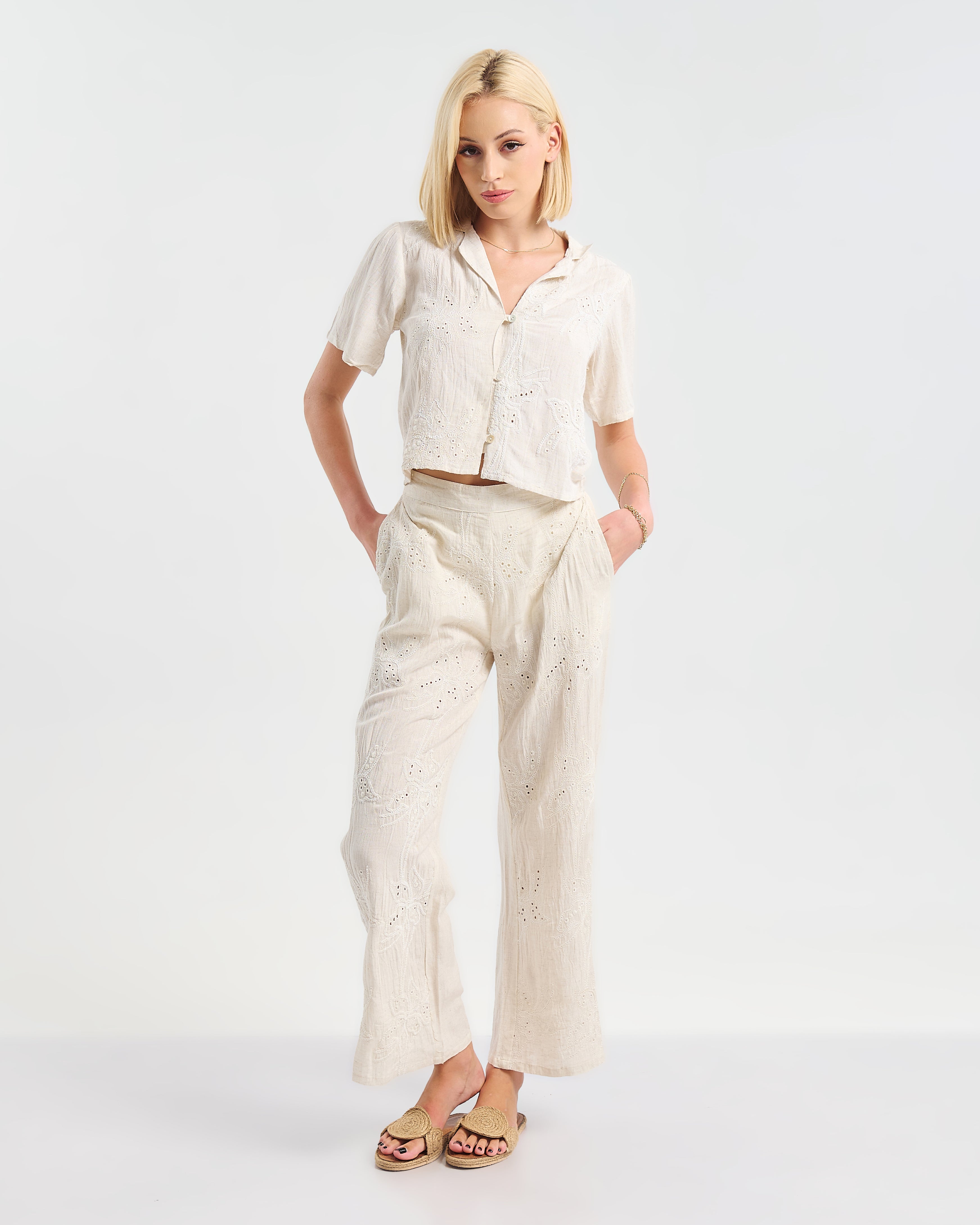 Model trägt ein cremefarbenes zweiteiliges Outfit aus kurzer Bluse mit Knopfleiste und weiter Hose. Beide Teile sind mit floraler Lochstickerei verziert. Kombiniert mit beigen Sandalen und dezentem Schmuck.“