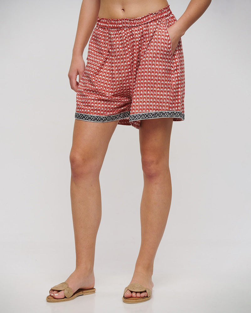 Nahaufnahme eines Models in locker fallenden Shorts mit elastischem Bund in Terrakotta und Weiß. Das feine, grafische Muster wird durch eine schwarze Bordüre mit ornamentalem Design am Saum ergänzt. Kombiniert mit beigen Sandalen.