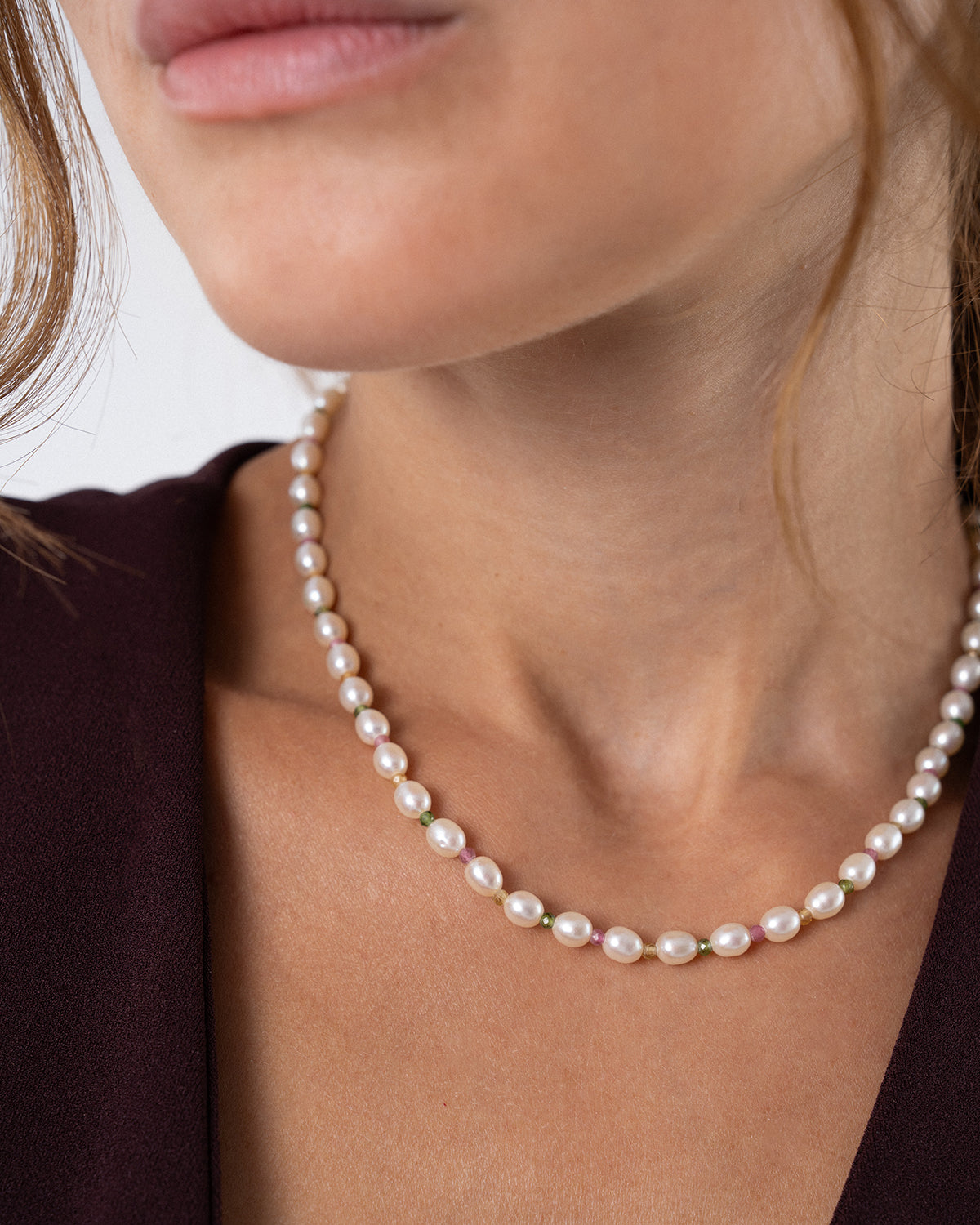 Detailaufnahme des Hals- und Dekolletébereichs einer Person mit einer feinen Perlenkette. Die Kette besteht aus kleinen, weißen Perlen, die von einzelnen farbigen Akzentperlen unterbrochen werden, und liegt auf einem dunkelbraunen Oberteil vor hellem Hintergrund.