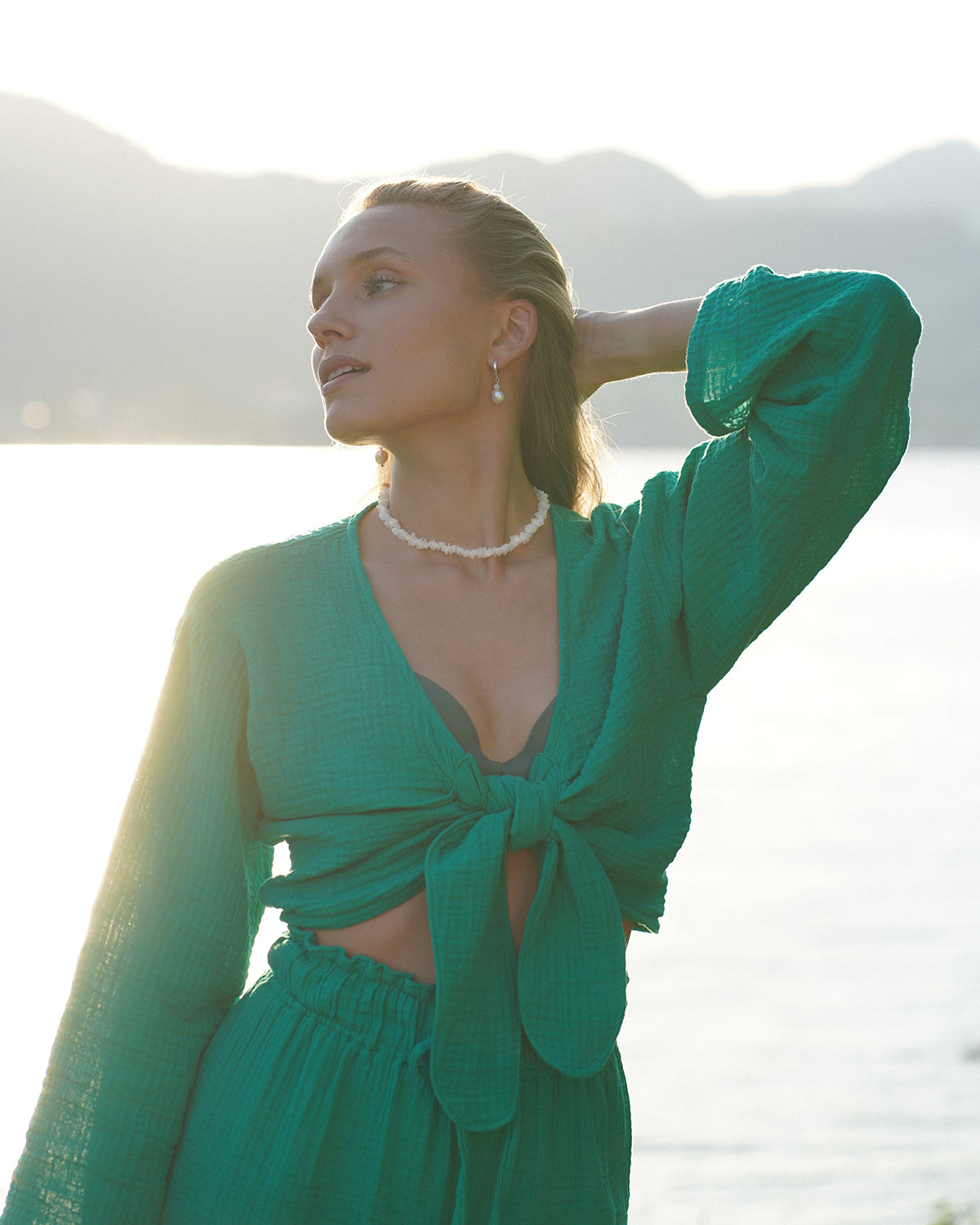 Frau im grünen Zweiteiler trägt eine weiße Mondstein-Halskette und passende Ohrhänger, aufgenommen im warmen Sonnenuntergang vor ruhiger See- und Bergkulisse im natürlichen Slow-Living-Stil.
