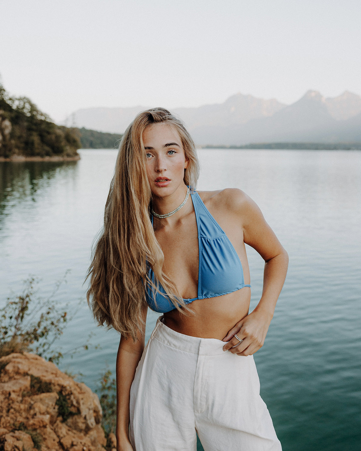 Junge Frau mit langen blonden Haaren trägt eine hellblaue Perlen-Choker-Halskette, kombiniert mit blauem Bikini-Top und weißer Hose, am See vor Bergkulisse.