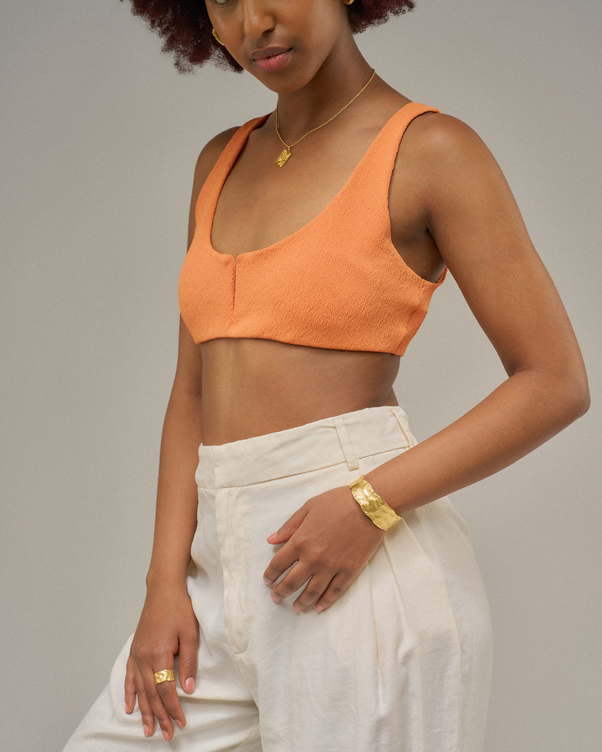 Nahaufnahme des Oberkörpers einer Person mit orangefarbenem Top und heller, hoch geschnittener Hose. Am Handgelenk trägt sie einen breiten, goldfarbenen Armreif; die Hand liegt locker an der Hüfte vor einem neutralen Hintergrund.