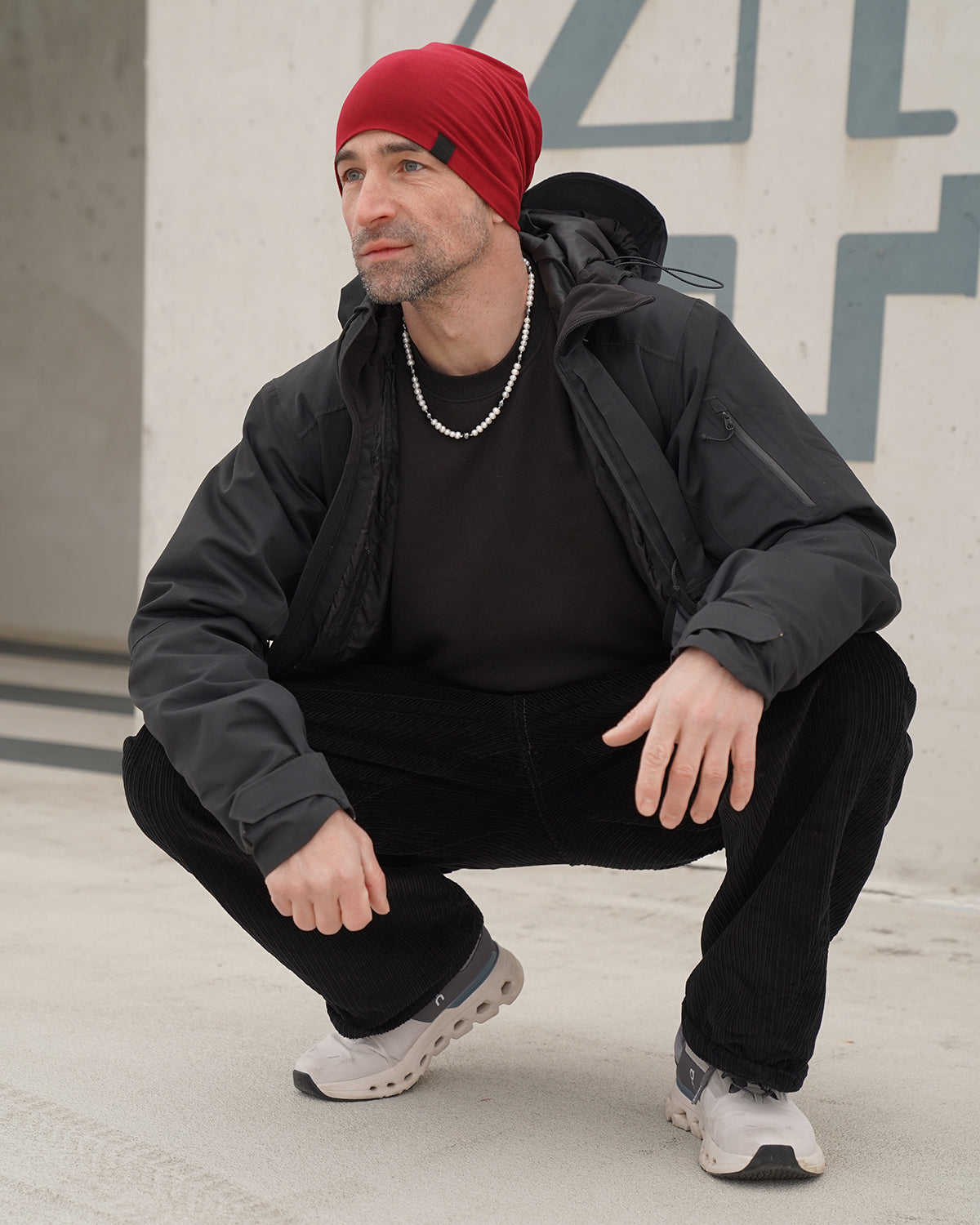Mann mit roter Mütze und schwarzer Outdoor-Jacke hockt im urbanen Umfeld und trägt eine Perlenkette für Herren aus weißen Perlen, sportlich-moderner Streetstyle.