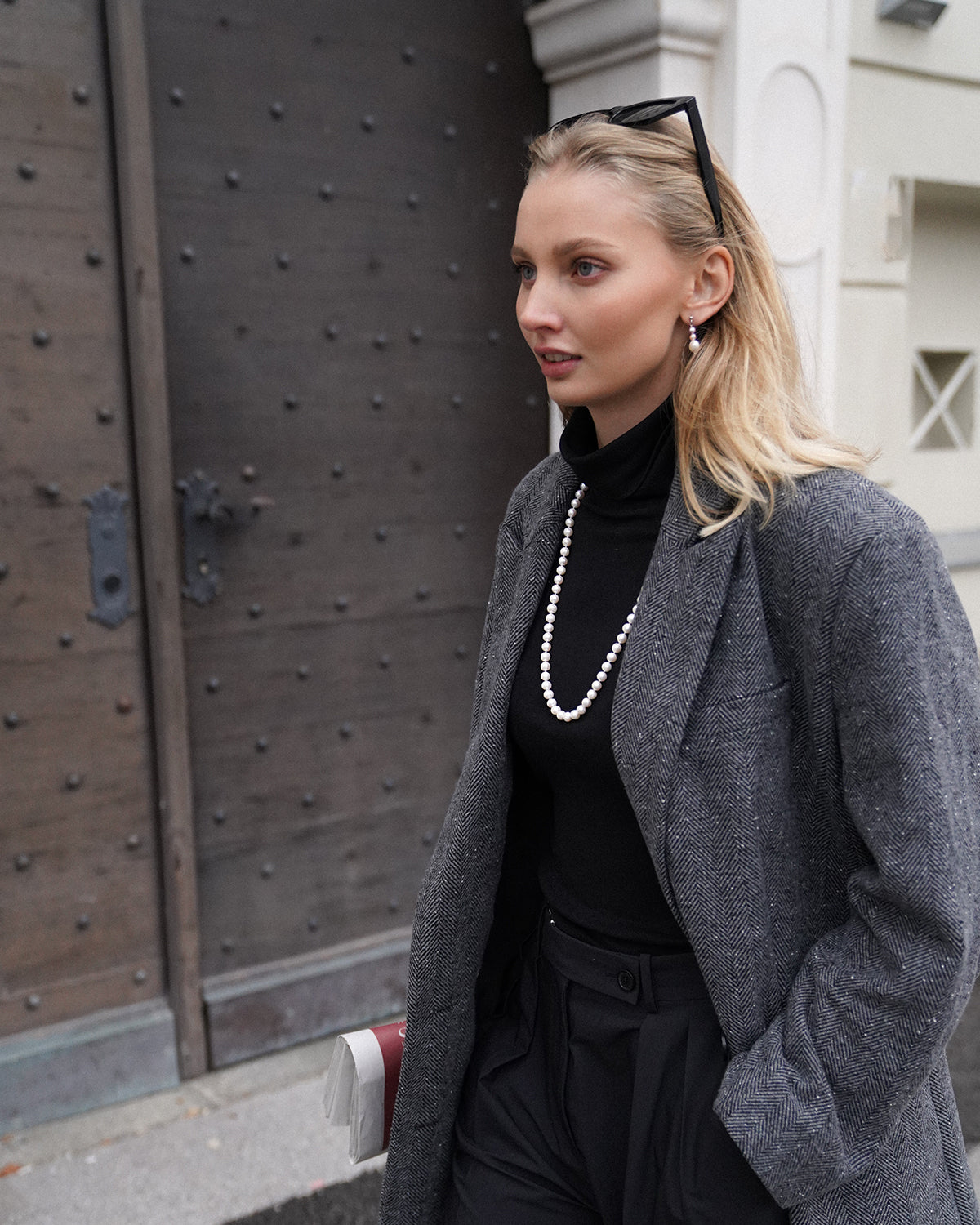 Seitliche Aufnahme einer blonden Frau vor einer großen Holztür, trägt schwarzen Rollkragenpullover, grauen Mantel im Fischgrätmuster und eine lange Perlenkette mit passenden Ohrringen; eine Sonnenbrille sitzt auf ihrem Kopf, unter dem Arm hält sie eine gefaltete Zeitung.