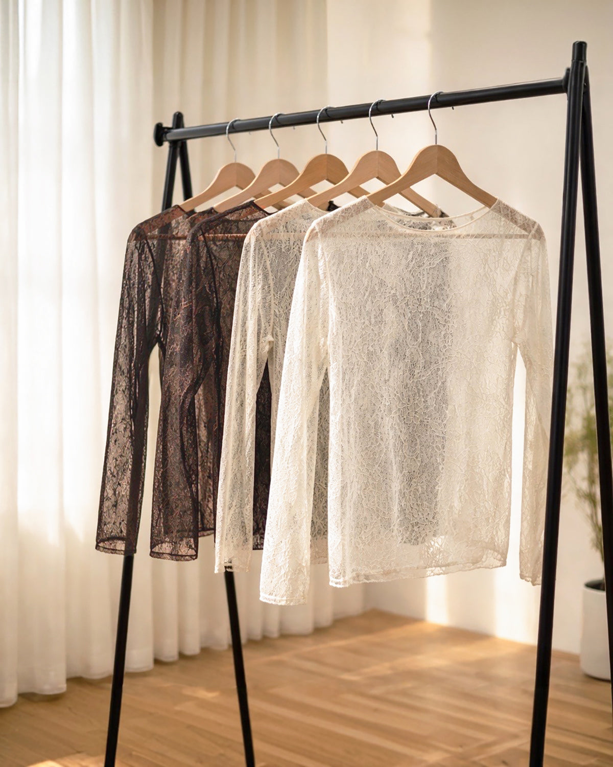 Mehrere langärmlige Spitzenoberteile in Schwarz, Dunkelbraun und Creme hängen auf Holzkleiderbügeln an einer schwarzen Kleiderstange in einem hellen Raum mit Vorhängen und Holzboden.