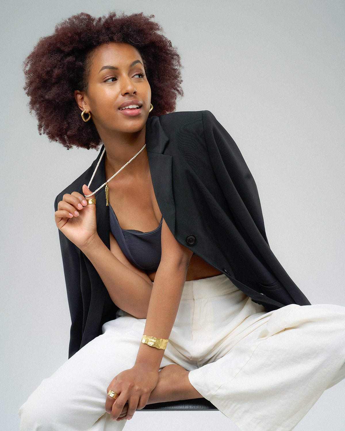 eine Frau die auf einem Sessel sitzt und  an ihrer Perlenkette mit goldenem Fischanhänger spielt. Der Hintergrund ist weiß.