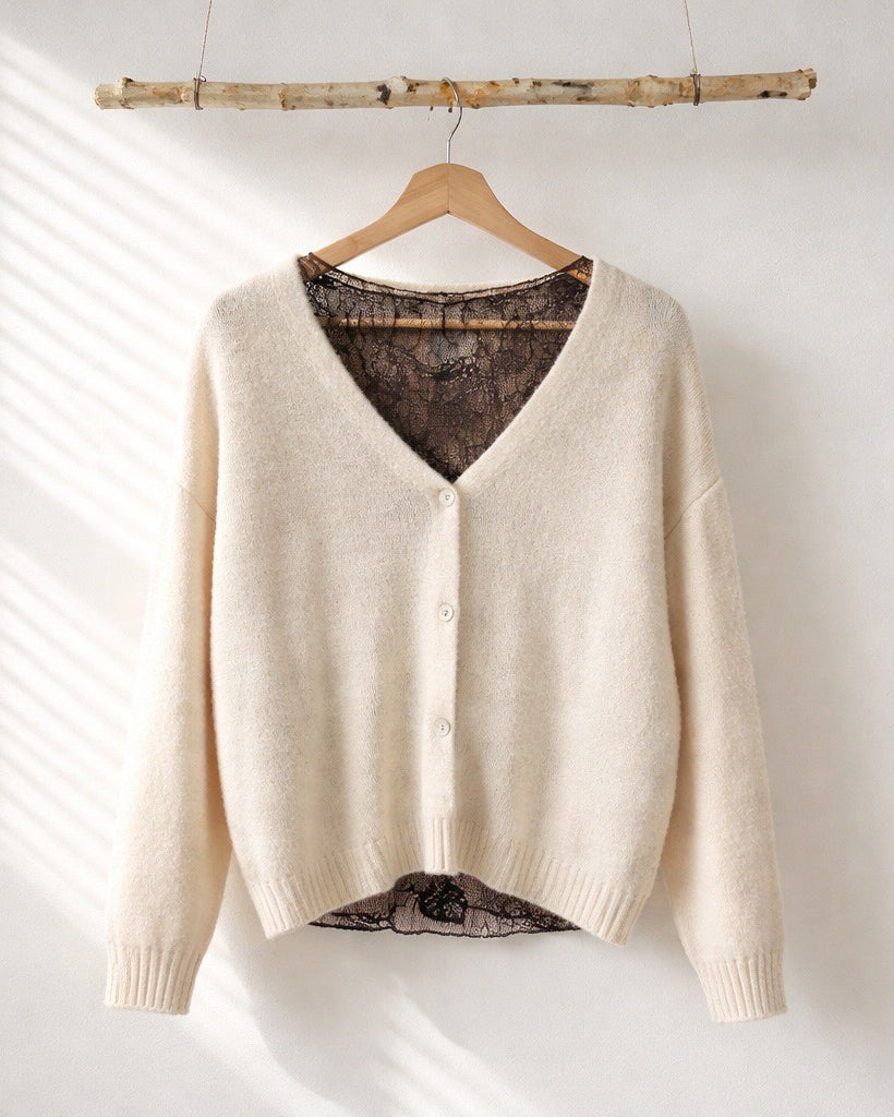 Beiger Strick-Cardigan mit V-Ausschnitt und Knöpfen, kombiniert mit braunem Spitzen-Top im Layering-Look, hängend an einem Holzkleiderbügel vor heller Wand.